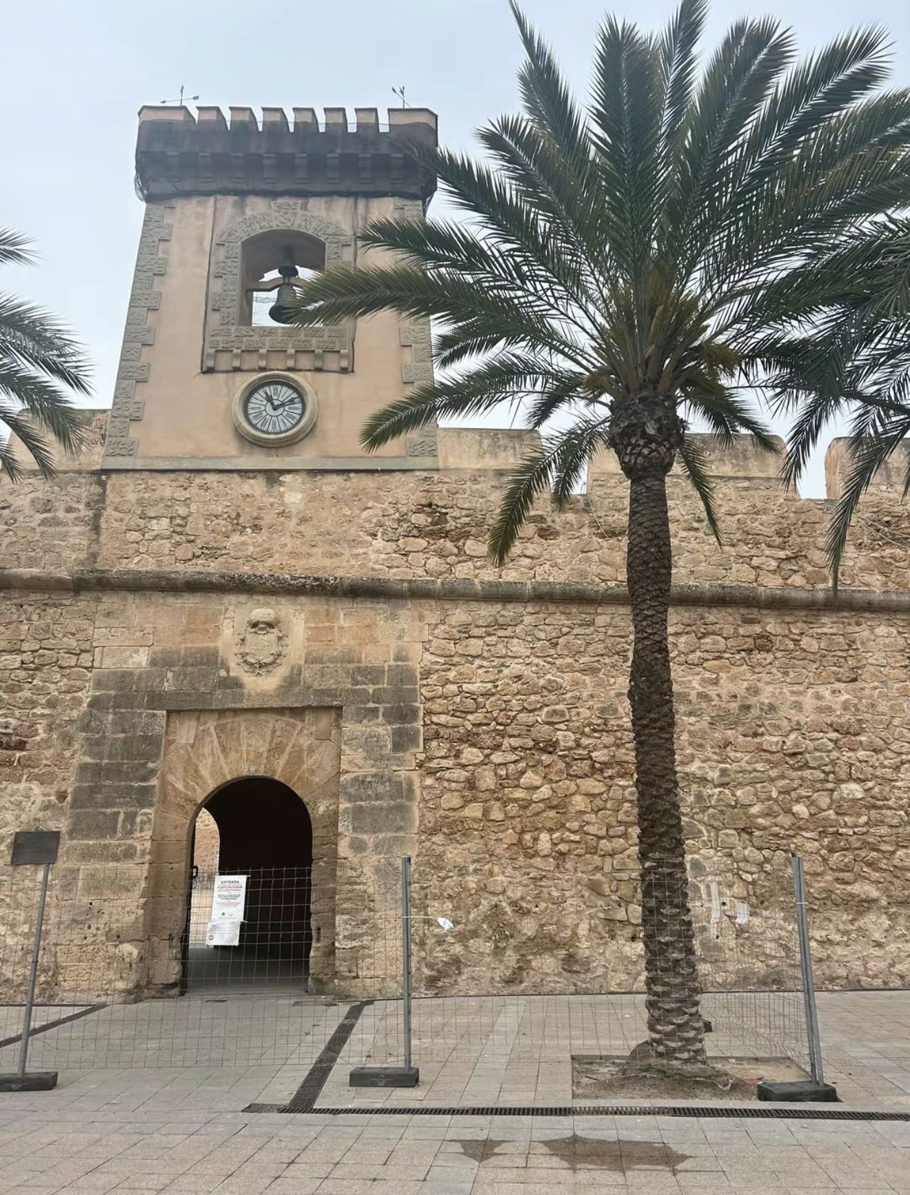 The clock tower and stone walls of the Castillo-Fortaleza in Santa Pola, framed by palm trees